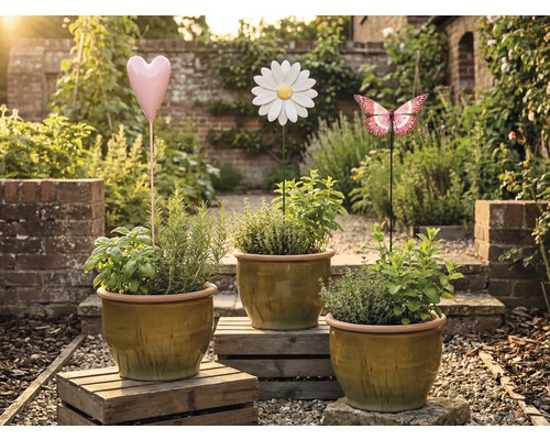 Drie plantenpotten met kruiden en decoratieve tuinpluggen in de vorm van een hart, bloem en vlinder in een tuin.