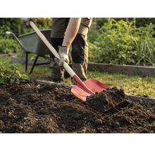 Een persoon spit met een spade de grond in de tuin om. Een kruiwagen staat op de achtergrond.