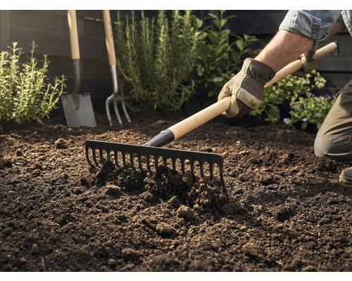 Tuinman harkt aarde met een hark, op de achtergrond zijn een spade en een riek te zien.