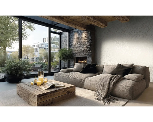 Lichte woonkamer met grijze bank, rustieke houten tafel, stenen haard en grote ramen. Wand met subtiel behang en houten plafondbalken.