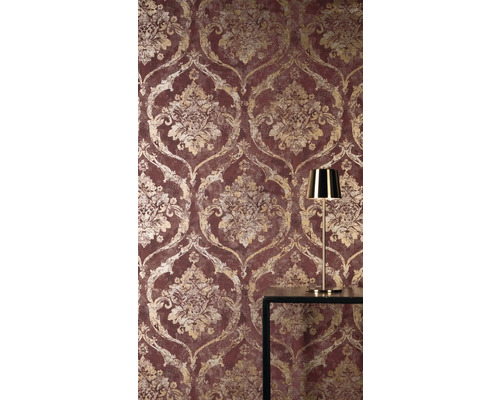 Vliesbehang met barok damastpatroon in goudlook op rode achtergrond, met een gouden tafellamp op een donkere tafel.