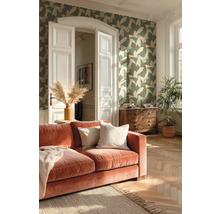 Woonkamer met vliesbehang met bladmotief, fluwelen bank, visgraatparket en houten ladekast in een lichte omgeving.