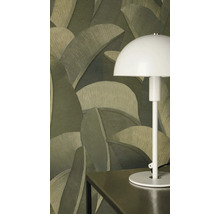 Vliesbehang met groot palmbladmotief in verschillende groentinten achter een witte tafellamp op een donkere tafel.