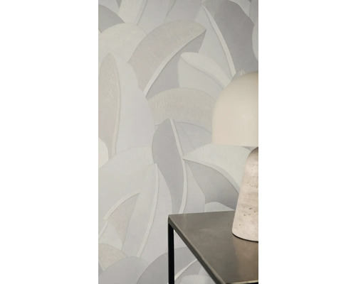 Vliesbehang met groot, gestructureerd bladpatroon in neutrale grijstinten op een muur achter een moderne tafellamp.