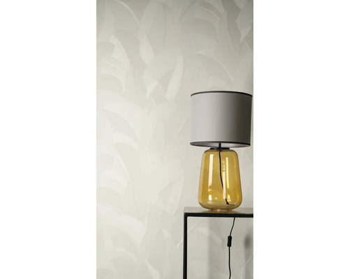 Vliesbehang met een subtiel bladerpatroon achter een tafellamp met gele glazen voet en grijze stoffen kap op een zwarte bijzettafel.