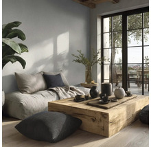 Moderne woonkamer met lichtgrijze muurverf, massief houten tafel, vloerkussens en grote raampartij naar het balkon.