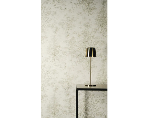 Vliesbehang met subtiel boompatroon en bosmotief in lichte tinten achter een gouden tafellamp op een zwarte tafel.