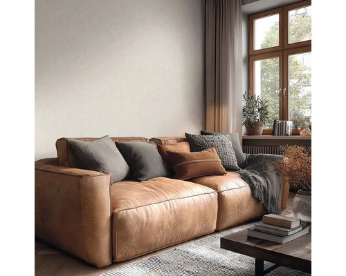 Lichte woonkamer met grote bruine leren bank, diverse kussens, grijze deken, behang met textuur en houten raam.