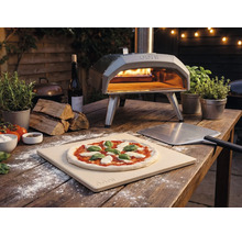 Buitenopname van een Ooni pizzaoven met pizza, pizzaschep en ingrediënten op een houten tafel.