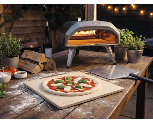 Buitenopname van een Ooni pizzaoven met pizza, pizzaschep en ingrediënten op een houten tafel.