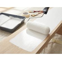 Houten plank wordt geverfd met een verfroller en witte verf, verfbak, penselen en afplakband liggen klaar.