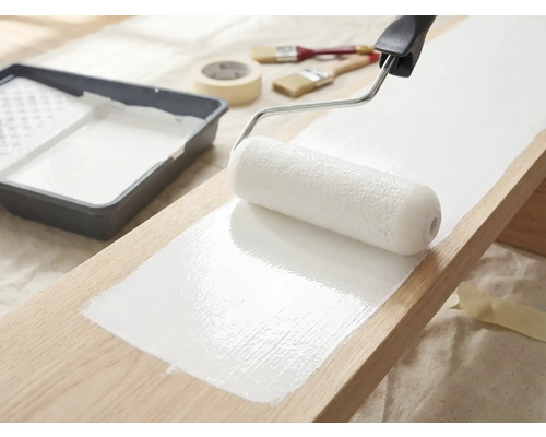 Houten plank wordt geverfd met een verfroller en witte verf, verfbak, penselen en afplakband liggen klaar.