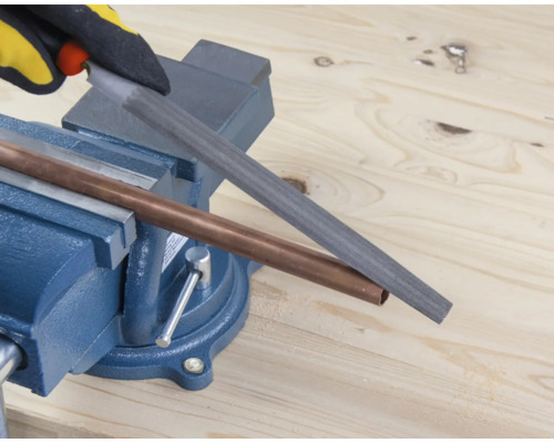 Een koperen buis wordt bewerkt met een vijl die in een bankschroef is geklemd.