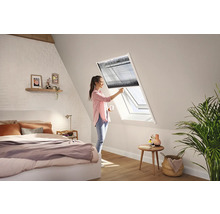 Een vrouw bedient een donker plisségordijn bij een wit dakraam in een lichte, moderne slaapkamer onder een schuin dak.