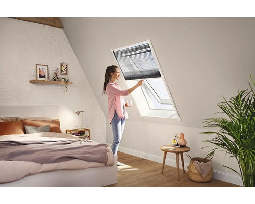 Een vrouw bedient een donker plisségordijn bij een wit dakraam in een lichte, moderne slaapkamer onder een schuin dak.