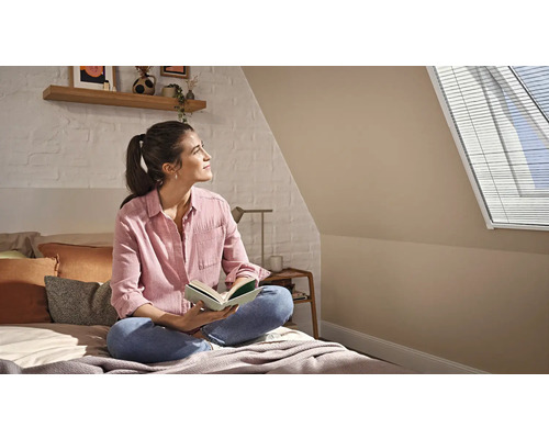 Vrouw leest op een bed in een lichte zolderkamer met dakraamjaloezie, witte bakstenen muur en houten plank.