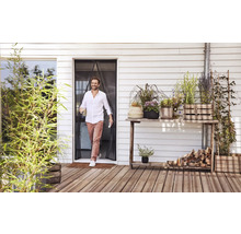 Een man loopt door een magnetisch insectengordijn bij een terrasdeur naar een houten terras met diverse potplanten.