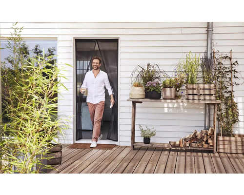 Een man loopt door een magnetisch insectengordijn bij een terrasdeur naar een houten terras met diverse potplanten.