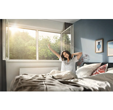 Lichte slaapkamer met een wijd openstaand kunststof raam waar zonlicht doorheen valt en een persoon in bed die zich bij het ontwaken uitrekt.