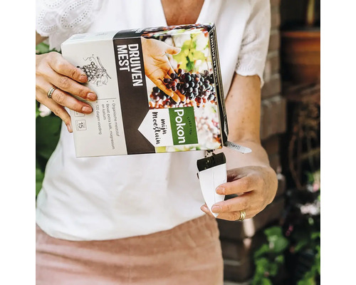 Een persoon giet Pokon biologische druivenmest uit een kartonnen verpakking in een witte maatbeker voor nauwkeurige dosering.