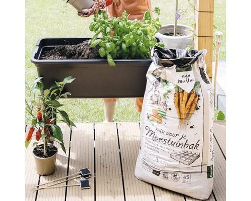 Aanplanten van een zwarte kweektafel naast een zak van 45 liter mix voor de moestuinbak op een houten vlonder.