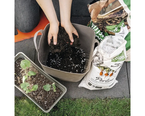 Iemand mengt potgrond met Perliet in een grijze kunststof teil. Ernaast staan zakken Pokon grond en Perliet en een tray met zaailingen.
