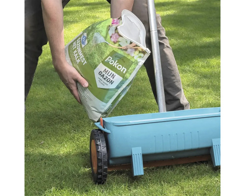 Een persoon vult een blauwe strooiwagen met Pokon Gazonmest met kalk vanuit een zak op een gazon.