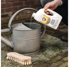 Home Defense Cure groene aanslagreiniger wordt vanuit een witte fles in een zinken gieter gegoten, met een houten borstel op de grond.