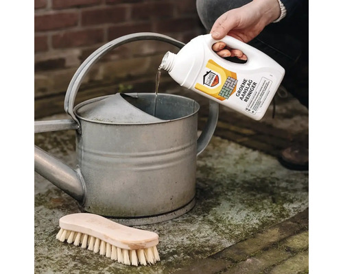 Home Defense Cure groene aanslagreiniger wordt vanuit een witte fles in een zinken gieter gegoten, met een houten borstel op de grond.