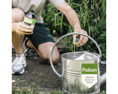 Een persoon giet vloeibare Pokon plantenvoeding met een maatbekertje in een metalen gieter in een tuin. Pokon Logo.