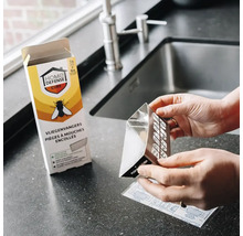 Handen houden een driehoekige vliegenvanger van Home Defense Cure vast bij een gootsteen met de verpakking op de achtergrond.