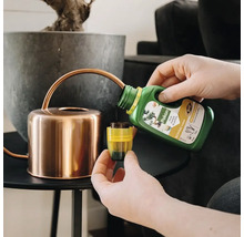 Een persoon schenkt vloeibare Pokon Bio Bonsai voeding uit een groene fles in een gele doseerdop naast een koperkleurige gieter.