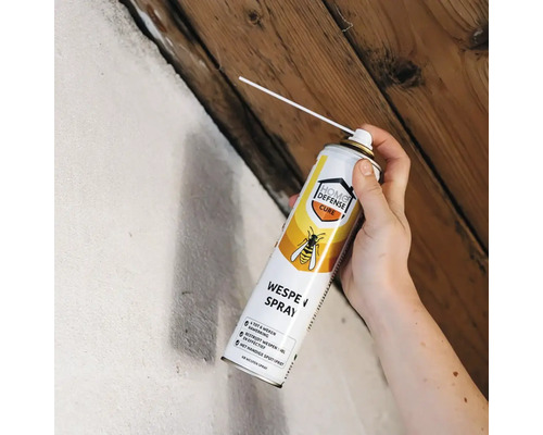 Hand spuit wespenspray met precisietuit in een kier tussen witte muur en houten plafond.