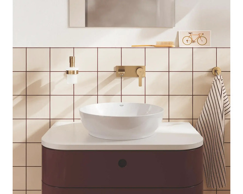 Heldere, georganiseerde badkamer met een witte opzetwastafel op een donkerrood meubel, gouden inbouwkranen en betegelde wanden.