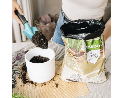 Een hand vult een witte geribbelde bloempot met Pokon kamerplanten potgrond met behulp van een schepje tijdens het verpotten.