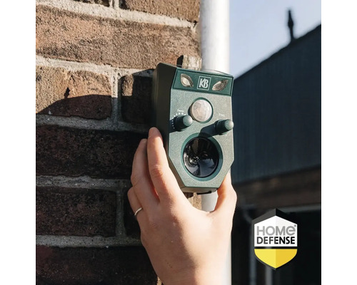 Een hand houdt een groene KB ultrasone dierenverjager tegen een bakstenen muur. Home Defense Logo.