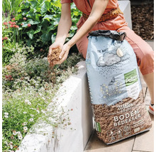 Een persoon strooit Pokon Water Proof bodembedekker uit een zak over planten in een lichte tuin.