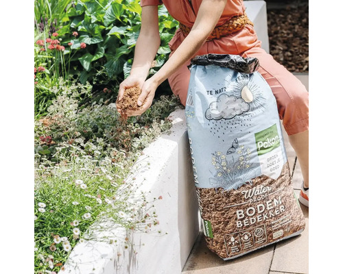 Een persoon strooit Pokon Water Proof bodembedekker uit een zak over planten in een lichte tuin.