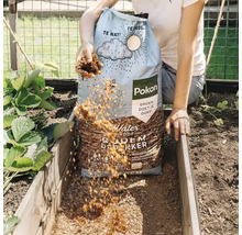 Een persoon strooit Pokon Waterproof Bodembedekker uit een 45 liter zak in een houten moestuinbak in de tuin naast aardbeienplanten.