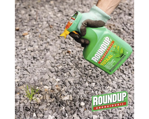 Een persoon met werkhandschoen spuit Roundup Onkruidvrij op onkruid tussen grind.