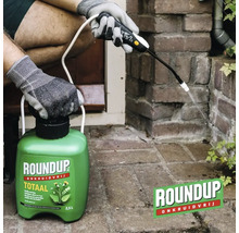 Een persoon met werkhandschoenen brengt Roundup Totaal onkruidverdelger aan met een 2,5 liter drukspuit op onkruid tussen de stenen.