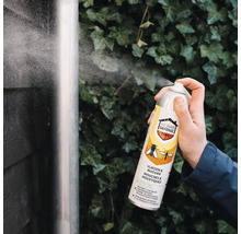 Een hand spuit insectenspray Home Defense Cure tegen vliegen en muggen op een regenpijp voor een wand met klimop.