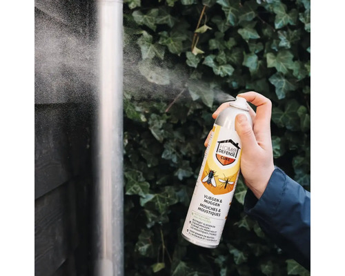 Een hand spuit insectenspray Home Defense Cure tegen vliegen en muggen op een regenpijp voor een wand met klimop.