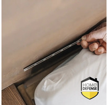 Een hand schuift een zilveren insectenwerende strip in een kier tussen matras en muur. Home Defense Logo.