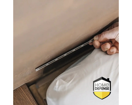 Een hand schuift een zilveren insectenwerende strip in een kier tussen matras en muur. Home Defense Logo.