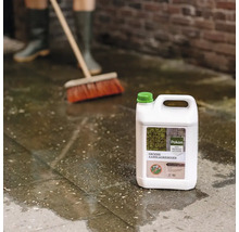 Pokon Groene Aanslagreiniger can op natte terrastegels, op de achtergrond wordt geveegd met een bezem.