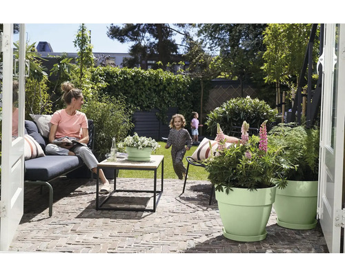 Modern terras met loungebank, houten tafel en lichtgroene bloempotten. Vrouw op de bank, kind spelend op het gazon in de achtergrond.
