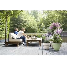 Lichte, ruime houten loungeset met lichte kussens op een grijs terras aan het water, versierd met plantenbakken en bloemen.