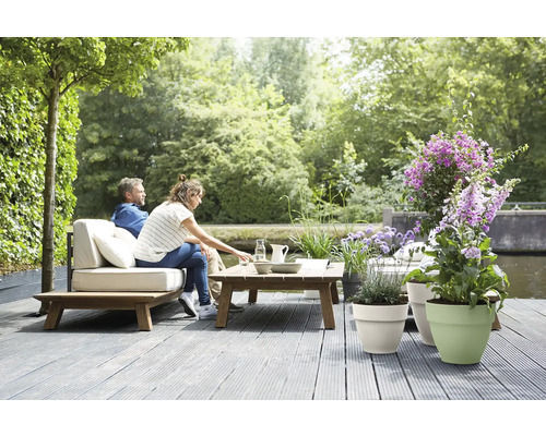 Lichte, ruime houten loungeset met lichte kussens op een grijs terras aan het water, versierd met plantenbakken en bloemen.