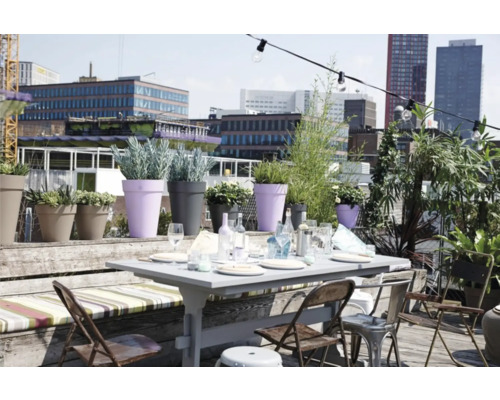 Licht dakterras met een gedekte tuintafel, klapstoelen, houten bank en diverse bloempotten tegen een stedelijke achtergrond.